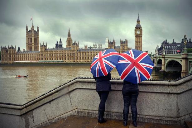 Zwei Personen mit Regenschirmen im Union-Jack-Design vor dem Palace of Westminster in London.