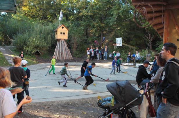 Kinder spielen mit Hockeyschlägern auf einem Spielplatz mit Baumhaus und Basketballkorb.