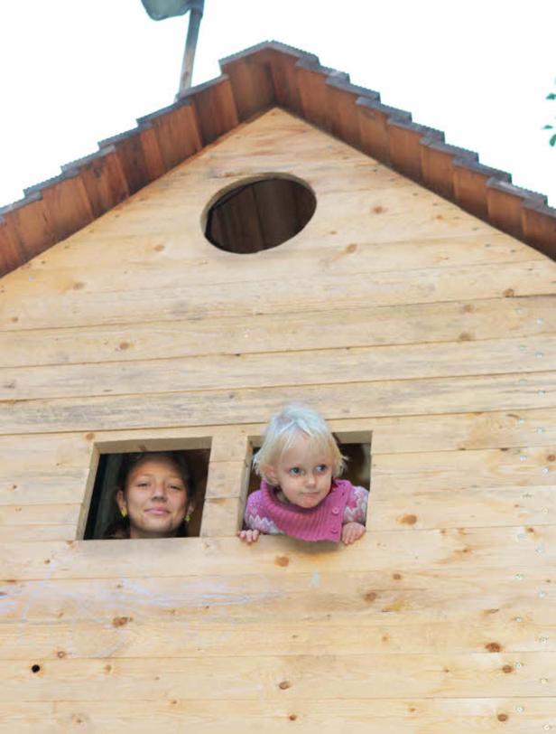 Zwei Mädchen schauen aus den Fenstern eines hölzernen Spielhauses.