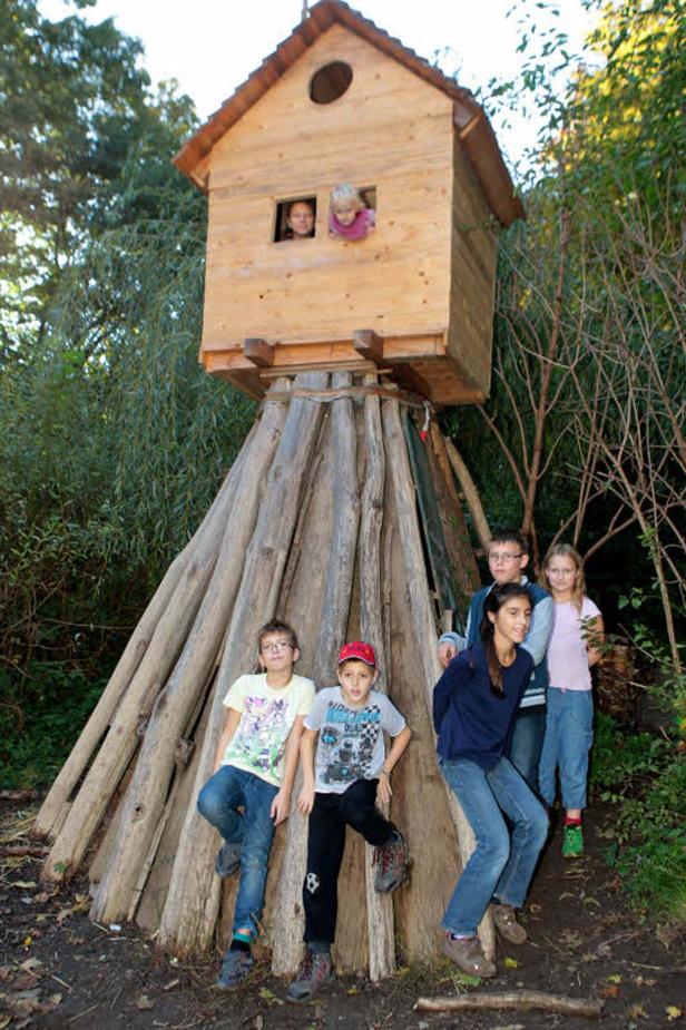 Eine Gruppe Kinder steht vor einem Baumhaus, zwei weitere schauen aus dem Fenster.