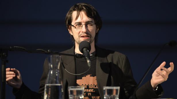 Ein Mann mit Brille spricht am Mikrofon vor einem dunklen Hintergrund.
