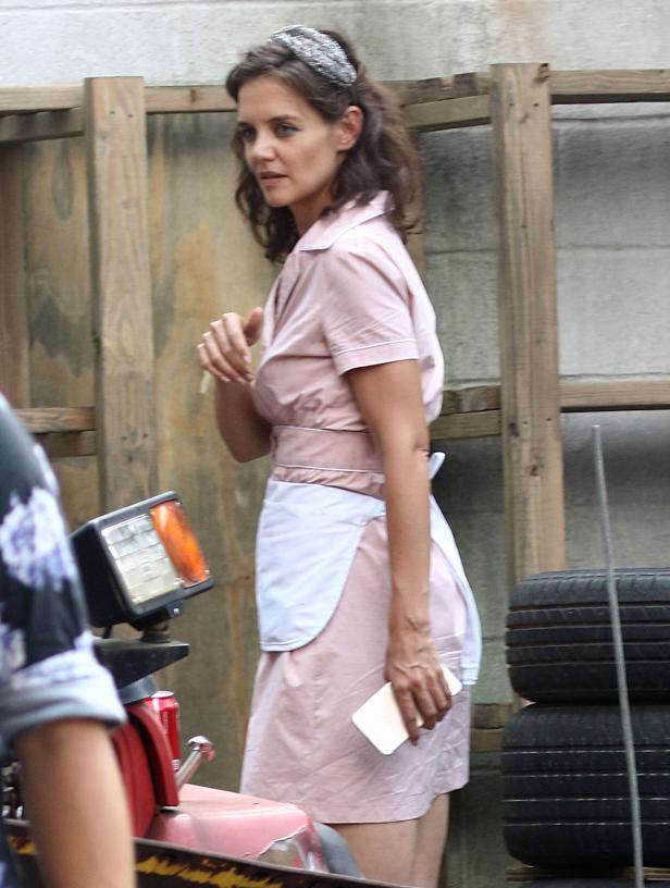 Katie Holmes in einem rosa Kleid mit weißer Schürze hält ein Handy in der Hand.
