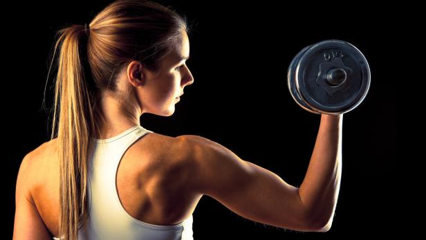 Eine Frau trainiert ihren Bizeps mit einer Hantel vor schwarzem Hintergrund.