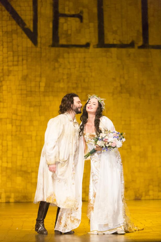 Ein Paar in historischen Kostümen steht auf einer Bühne vor einer gelben Wand.