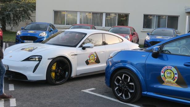 Mehrere Autos, darunter ein weißer Porsche und blaue Ford Pumas, stehen auf einem Parkplatz.