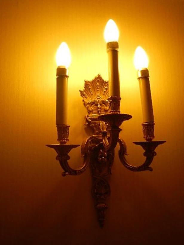 Eine goldene Wandleuchte mit drei brennenden Kerzen an einer gelben Wand.