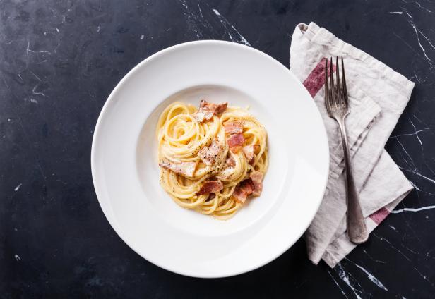 Ein Teller Spaghetti Carbonara mit Speckwürfeln und Pfeffer auf einem dunklen Tisch.