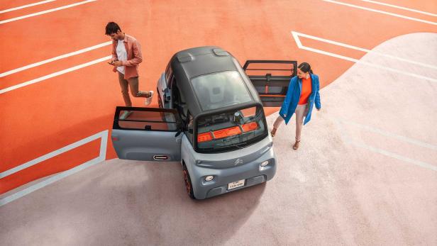 Ein grauer Citroën Ami parkt auf einer orangefarbenen Fläche, während zwei Personen einsteigen.