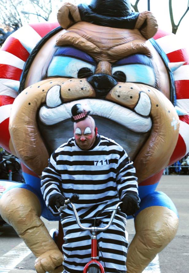 Ein als Sträfling verkleideter Clown fährt vor einem riesigen Bulldoggen-Kopf auf einem kleinen Fahrrad.