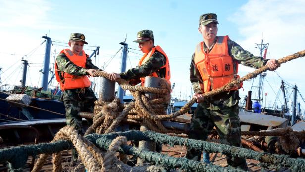 Drei Soldaten in Uniform und Schwimmwesten hantieren mit Seilen auf einem Schiff.