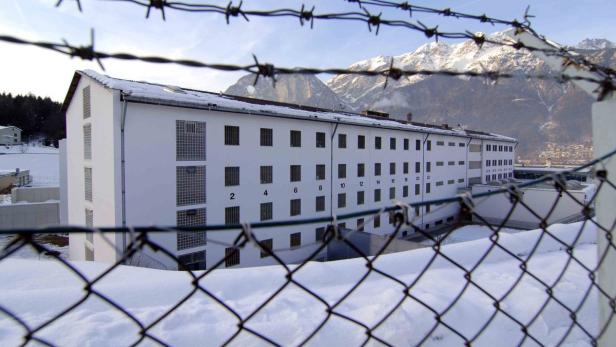 Ein weißes Gebäude mit vergitterten Fenstern und Stacheldraht im Vordergrund, im Hintergrund schneebedeckte Berge.