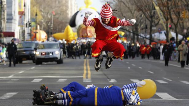 Ein Clown springt auf Rollschuhen über einen am Boden liegenden Clown bei einer Parade.