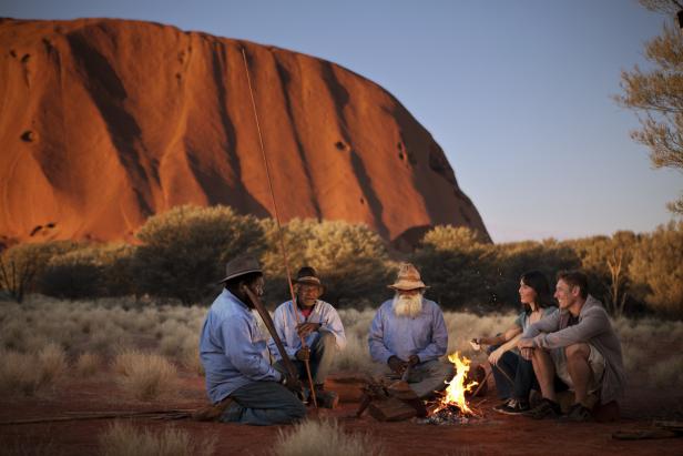 Eine Gruppe von Menschen sitzt am Lagerfeuer vor dem Uluru (Ayers Rock) in Australien.