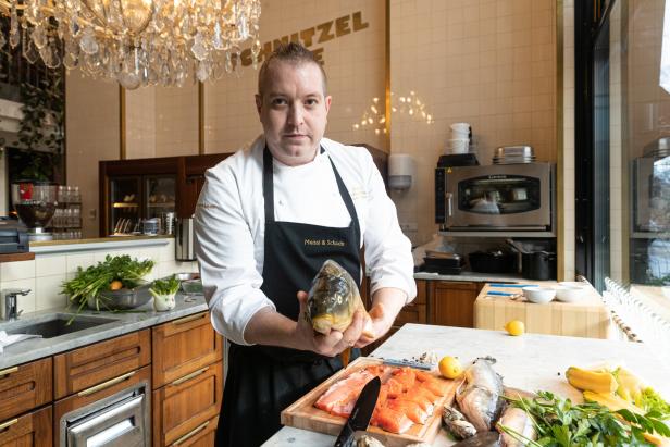 Ein Koch hält einen Karpfen in den Händen, während auf dem Tisch Fisch und Gemüse liegen.