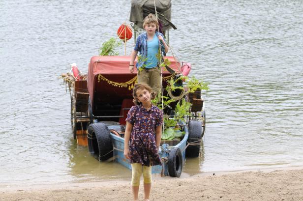 Ein Junge und ein Mädchen stehen vor einem selbstgebauten Boot am Ufer.