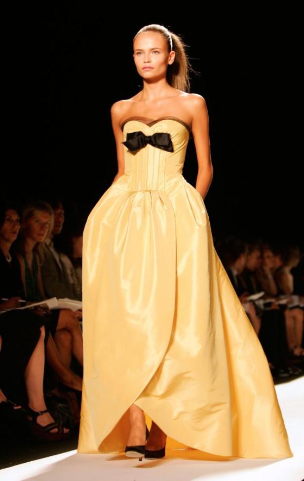 Ein Model präsentiert ein gelbes, trägerloses Abendkleid mit schwarzer Schleife auf dem Laufsteg.