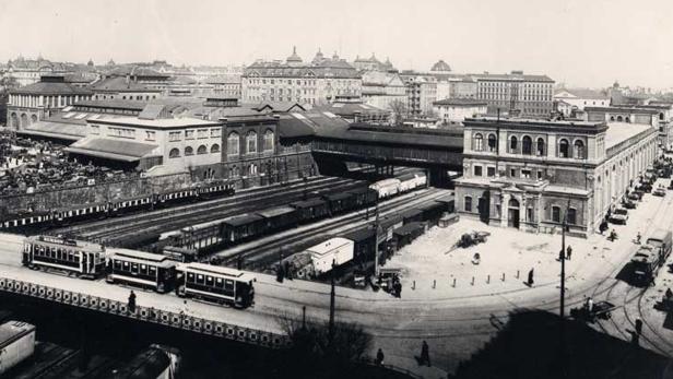 Historische Aufnahme eines belebten Bahnhofs mit Straßenbahnen und Zügen.