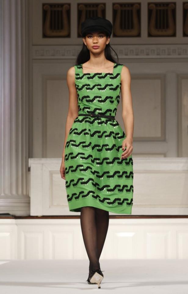 Eine Frau mit Hut präsentiert ein grünes Kleid mit schwarzen Mustern auf einem Laufsteg.