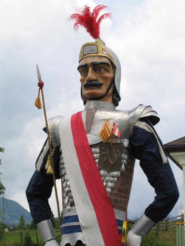 Eine große Ritterfigur mit Helm, Lanze und rot-weißem Band steht im Freien.