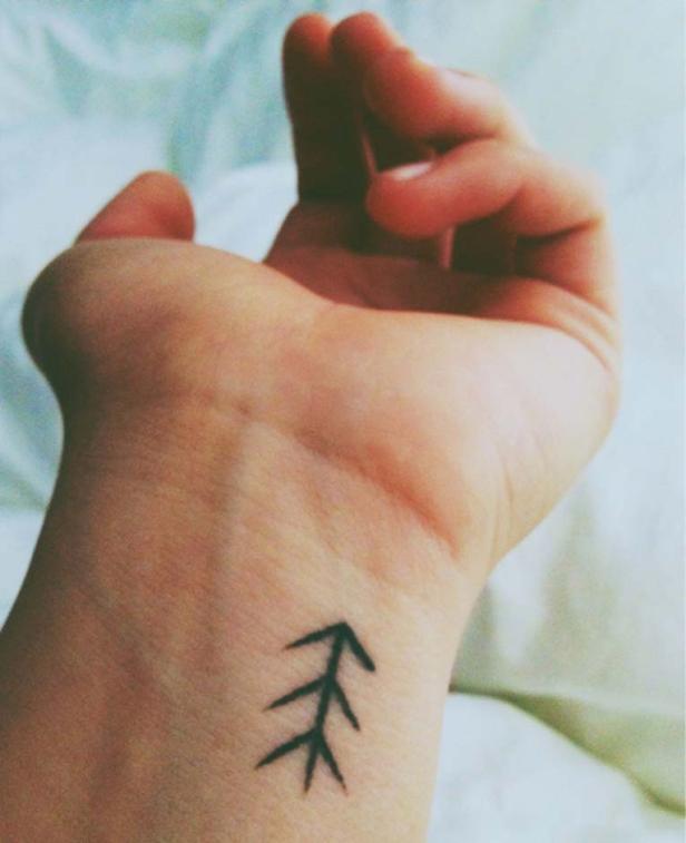 Auf dem Handgelenk einer Person befindet sich ein kleines, schwarzes Tattoo, das einen Baum darstellt.