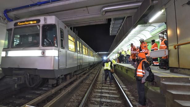 Eine U-Bahn steht an einem Bahnsteig, während Arbeiter in Warnwesten daneben stehen.