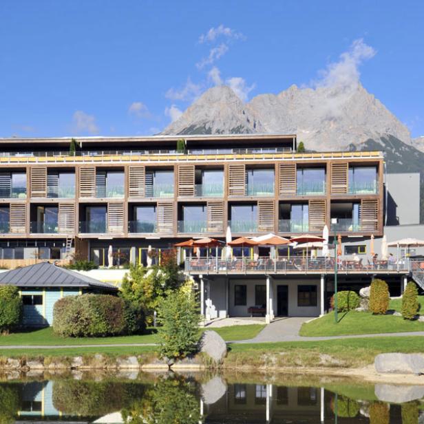 Ein modernes Hotel liegt vor einer Bergkulisse und einem kleinen See.