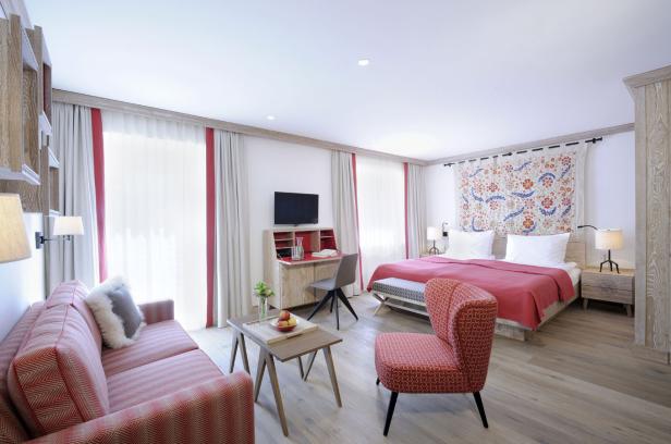 Ein helles Hotelzimmer mit Sofa, Sessel, Schreibtisch und einem Doppelbett mit roter Tagesdecke.