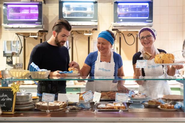 In der „Vollpension“ wählt ein Kunde Kuchen aus der Auslage, bedient von zwei freundlichen Verkäuferinnen.
