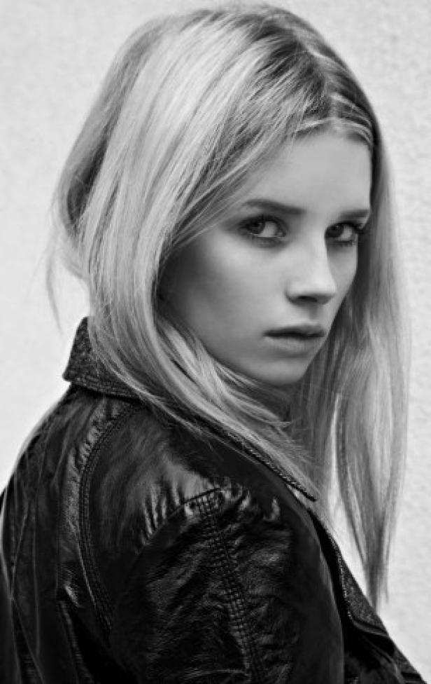 Eine junge Frau mit blonden Haaren und Lederjacke blickt über ihre Schulter.