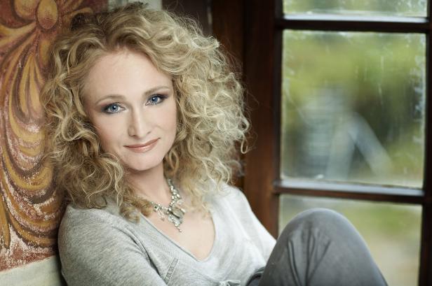 Eine Frau mit blonden Locken und blauem Blick lehnt an einer Wand neben einem Fenster.