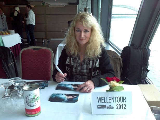 Blonde Frau sitzt an einem Tisch und signiert Autogrammkarten für die „Wellentour 2012“.