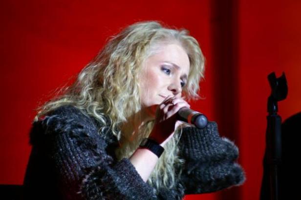 Eine Frau mit blonden Locken singt in ein Mikrofon vor einem roten Hintergrund.