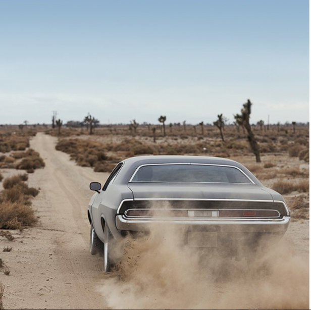 Ein grauer Oldtimer fährt auf einer unbefestigten Straße durch eine Wüstenlandschaft.