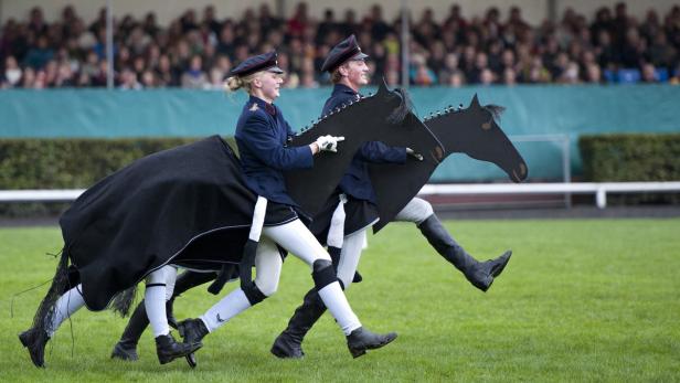 Zwei Personen in Uniformen führen eine Pantomime mit einem Pferde-Requisit auf einer Wiese auf.