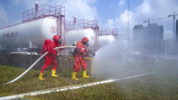 Zwei Arbeiter in Schutzanzügen sprühen Wasser auf Tanks in einer Industrieanlage.
