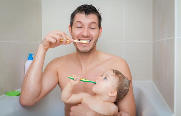 Ein Vater putzt sich die Zähne, während er sein Kind im Arm hält, das ebenfalls Zähne putzt.