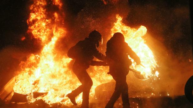 Zwei Personen stehen vor einem großen Feuer, möglicherweise bei einer Demonstration.