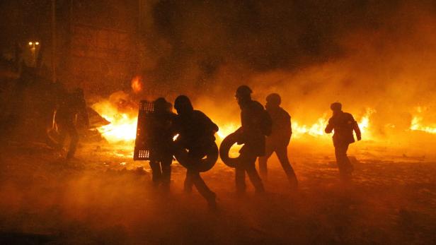 Mehrere Personen in der Dunkelheit, einige mit Schilden und Reifen, vor einem großen Feuer.