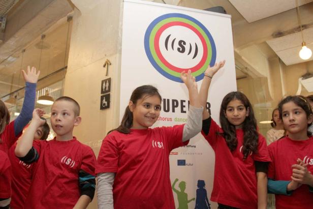 Eine Gruppe Kinder in roten T-Shirts steht vor einem Banner mit der Aufschrift „Superar Chorus Dance“.