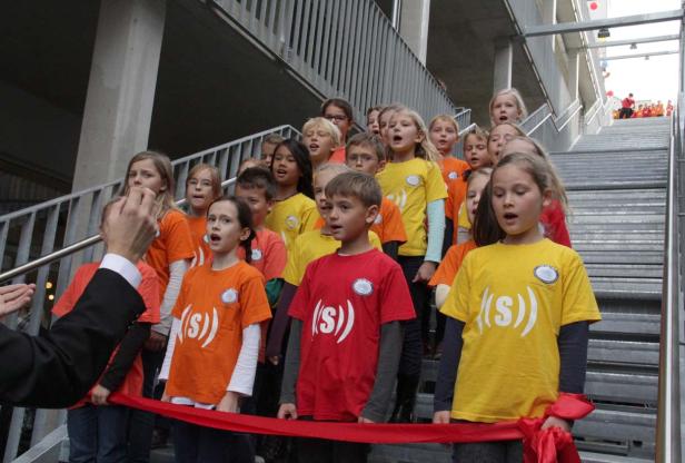Ein Kinderchor steht auf einer Treppe und singt bei einer Veranstaltung mit rotem Band.