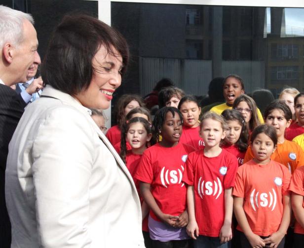 Eine Frau in einem hellen Blazer steht vor einer Gruppe singender Kinder.