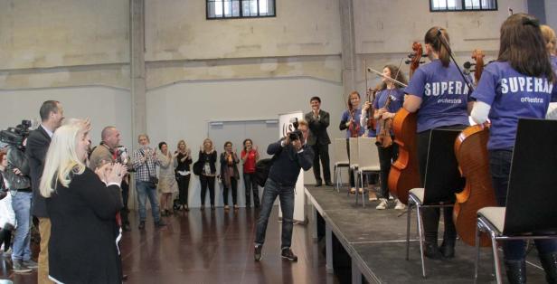Ein Orchester spielt vor Publikum, das applaudiert; ein Fotograf macht Fotos.