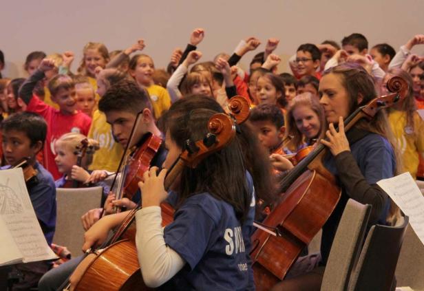 Ein Orchester von Kindern und Jugendlichen spielt Geige und Cello, während andere im Hintergrund jubeln.