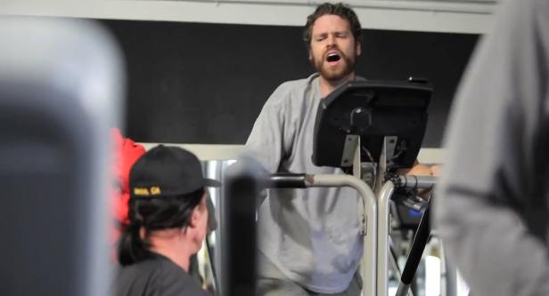 Ein Mann mit Bart trainiert erschöpft auf einem Laufband in einem Fitnessstudio.