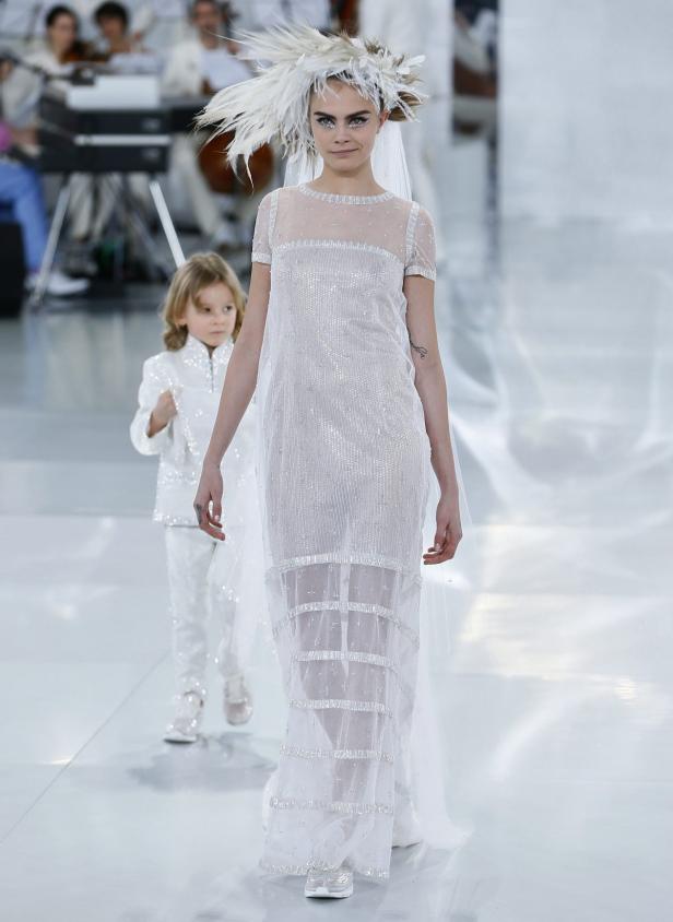 Cara Delevingne in einem weißen Kleid mit Federschmuck auf dem Laufsteg, begleitet von einem Kind.