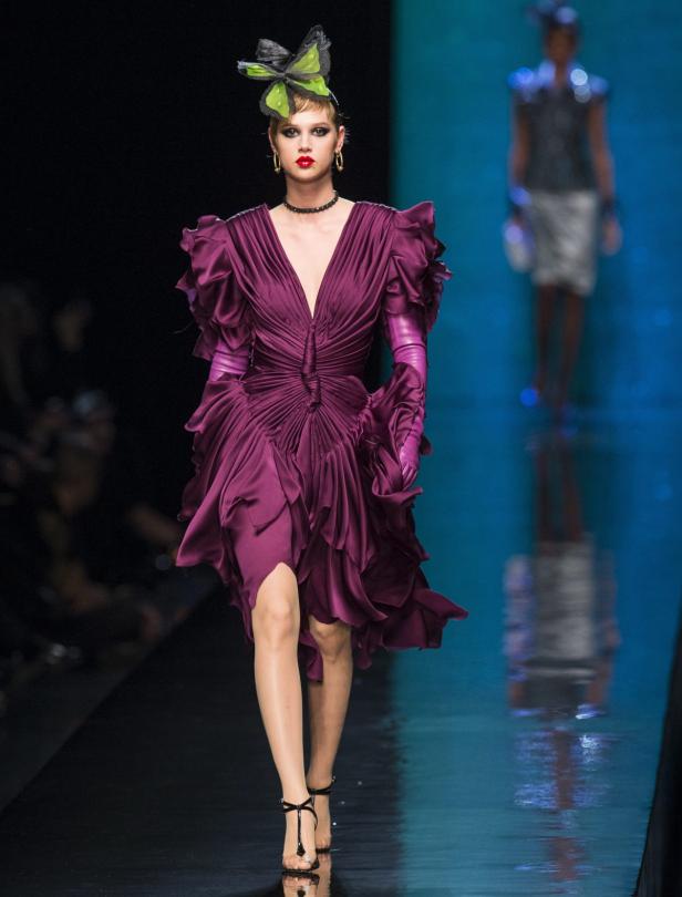 Eine Frau in einem violetten Kleid mit Rüschen und einem auffälligen Kopfschmuck auf dem Laufsteg.