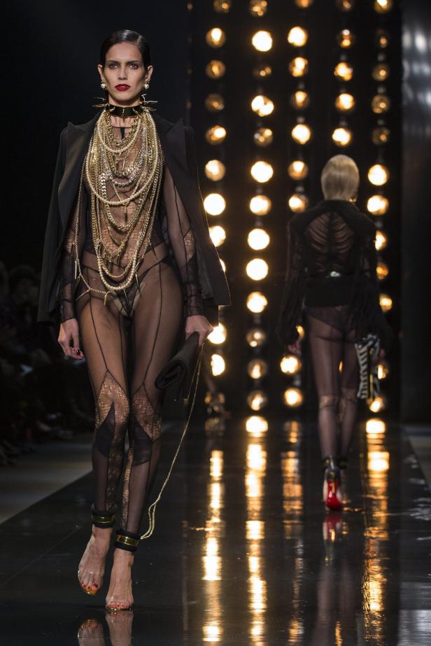 Eine Frau in durchsichtiger Kleidung und vielen goldenen Ketten auf einem Laufsteg.