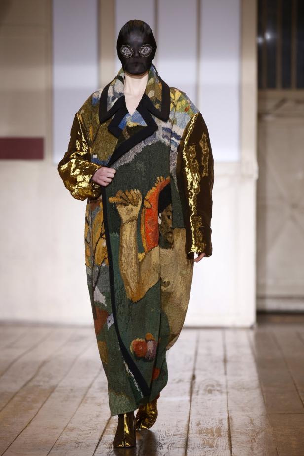 Ein Model mit einer schwarzen Maske und einem langen, gemusterten Mantel auf dem Laufsteg.