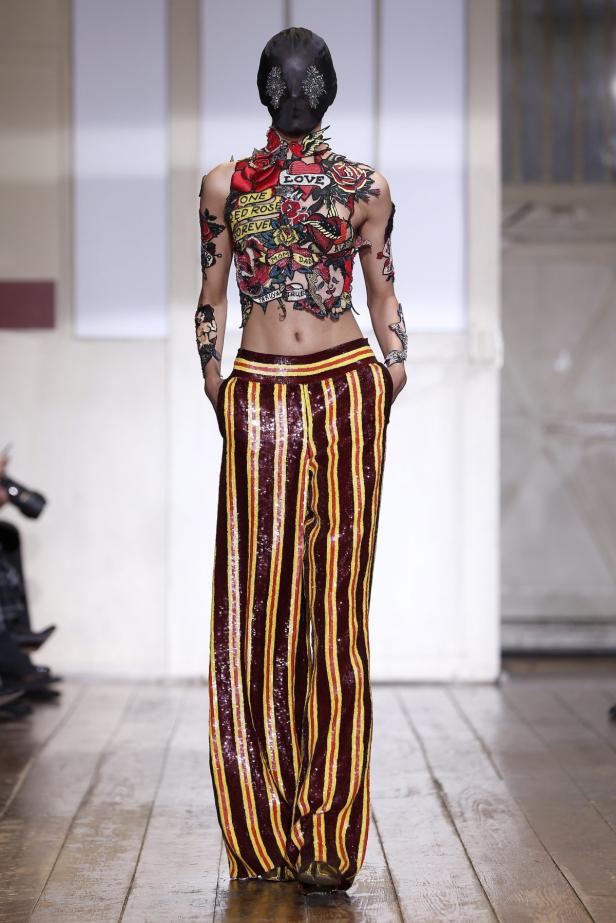 Ein Model mit einer schwarzen Gesichtsmaske und einem Oberteil mit Tattoo-Motiven und gestreiften Paillettenhosen.