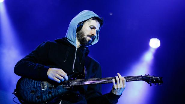 Ein bärtiger Mann mit Kapuzenpullover spielt E-Gitarre auf einer Bühne mit blauem Licht.